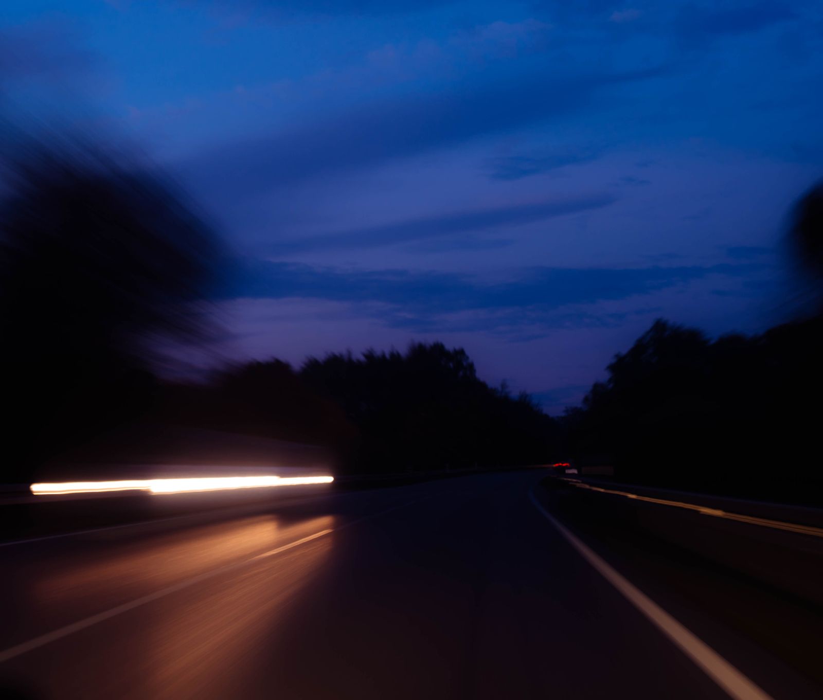 Nächtliche Landstraße in Bewegungsunschärfe, eine helle Lichtspur zieht vorbei, darüber tiefblauer Abendhimmel